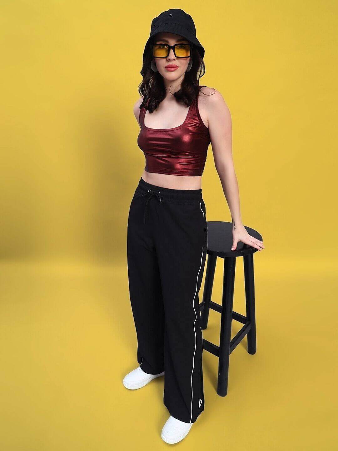 Woman modeling maroon-black co-ord set with metallic top and polyester joggers, wearing a hat and sunglasses against a yellow background.
