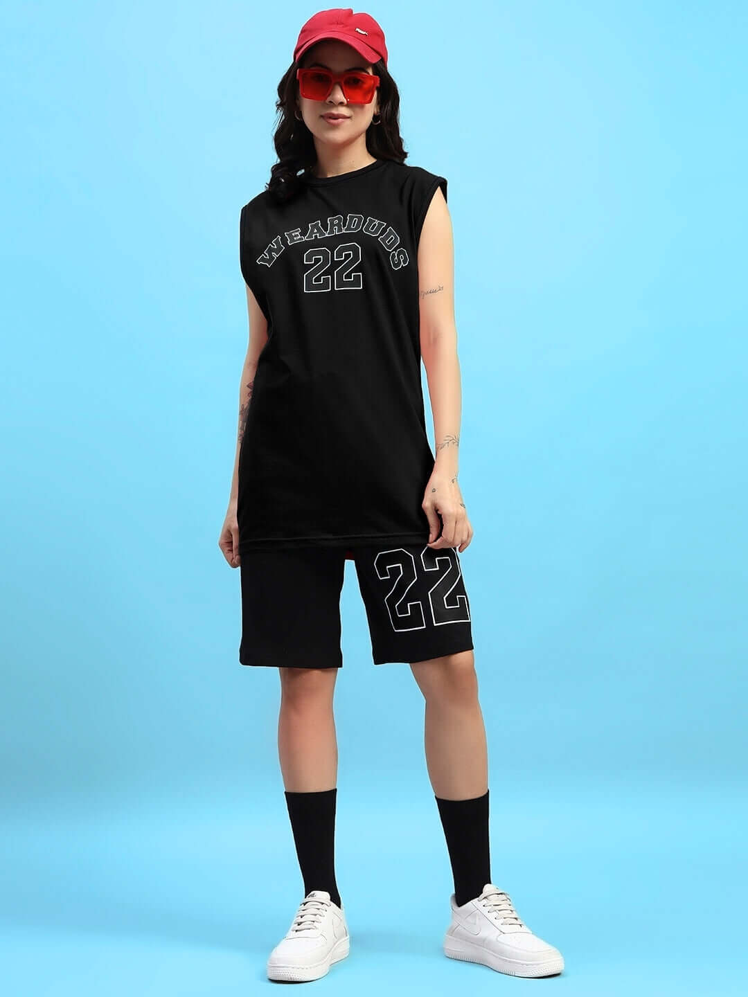 Woman wearing black gym co-ord set with number 22, red cap, sunglasses, standing on light blue background.