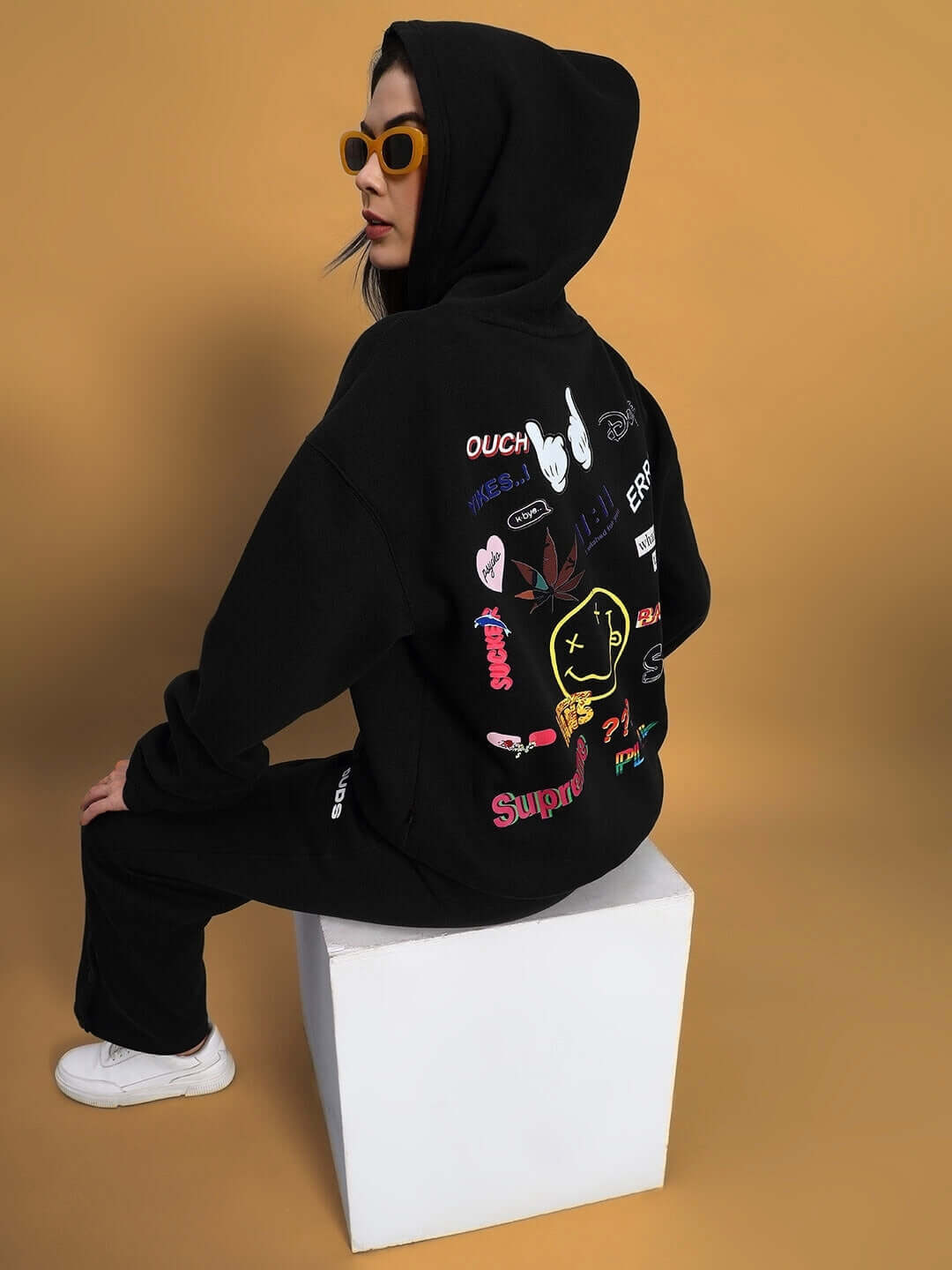 Woman wearing WOMEN'S SOOTY FLEECE CARGO PANT BLACK with graphic hoodie, sitting on a white cube, product display