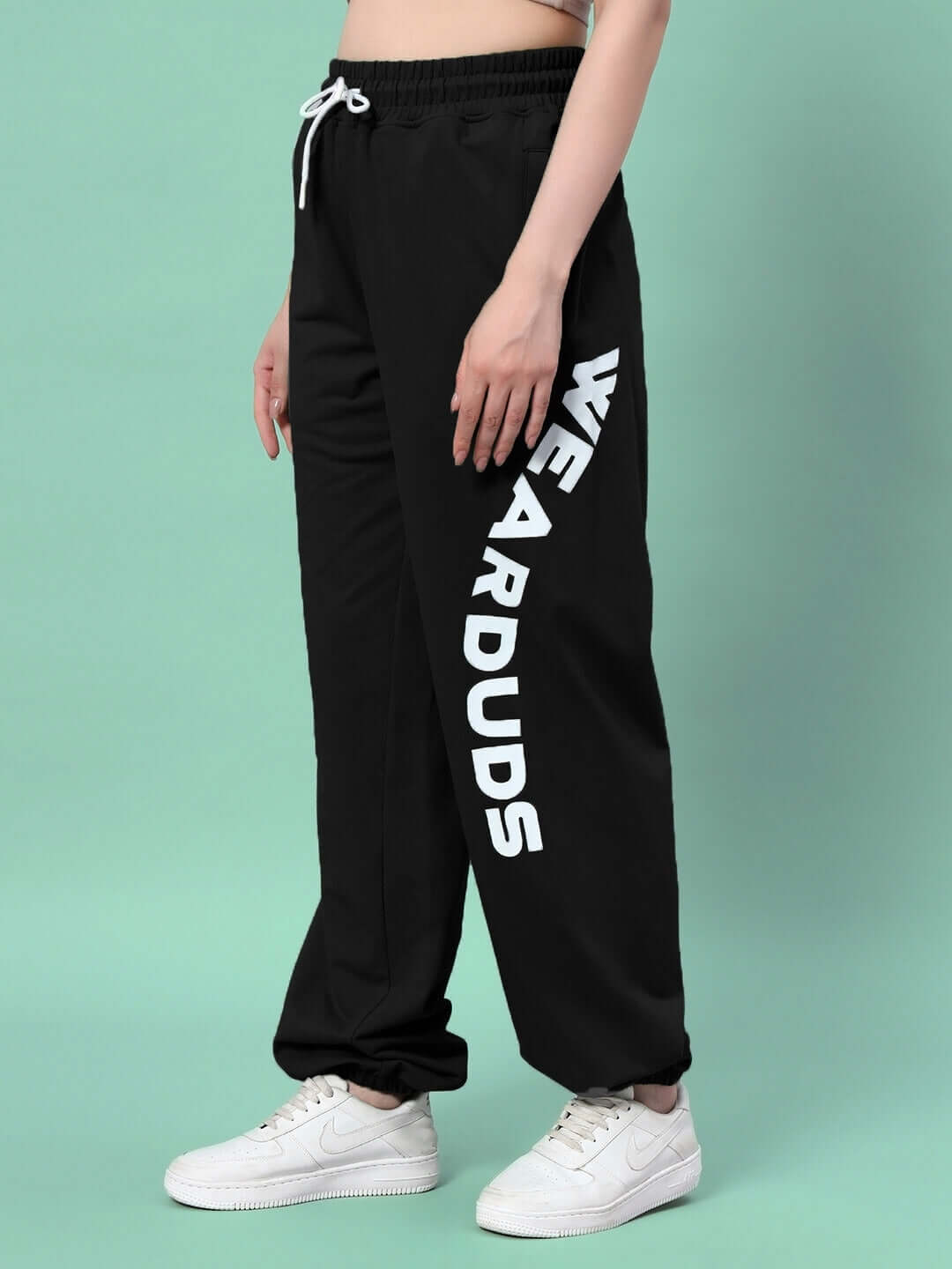 Model wearing WOMEN'S ELLEN RELAXED FIT JOGGER BLACK with white lettering on the leg, paired with white sneakers on a turquoise background.