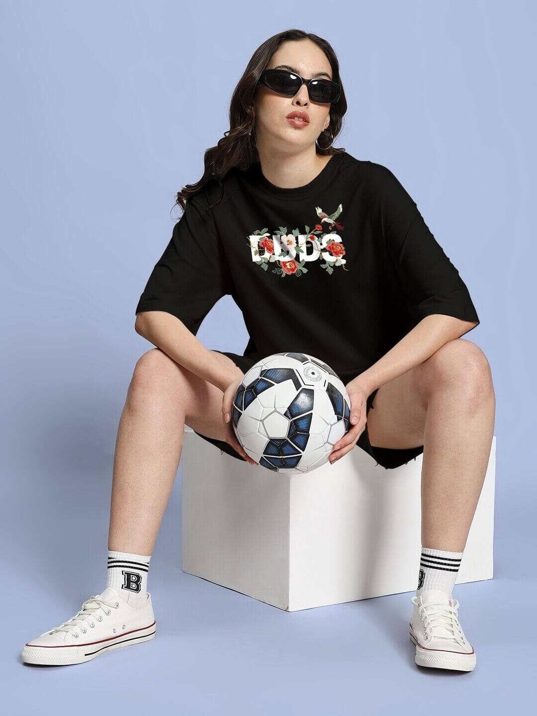 Woman wearing WOMEN ROSE CHARM OVER-SIZED T-SHIRT BLACK holding a soccer ball, showcasing chic streetwear style.