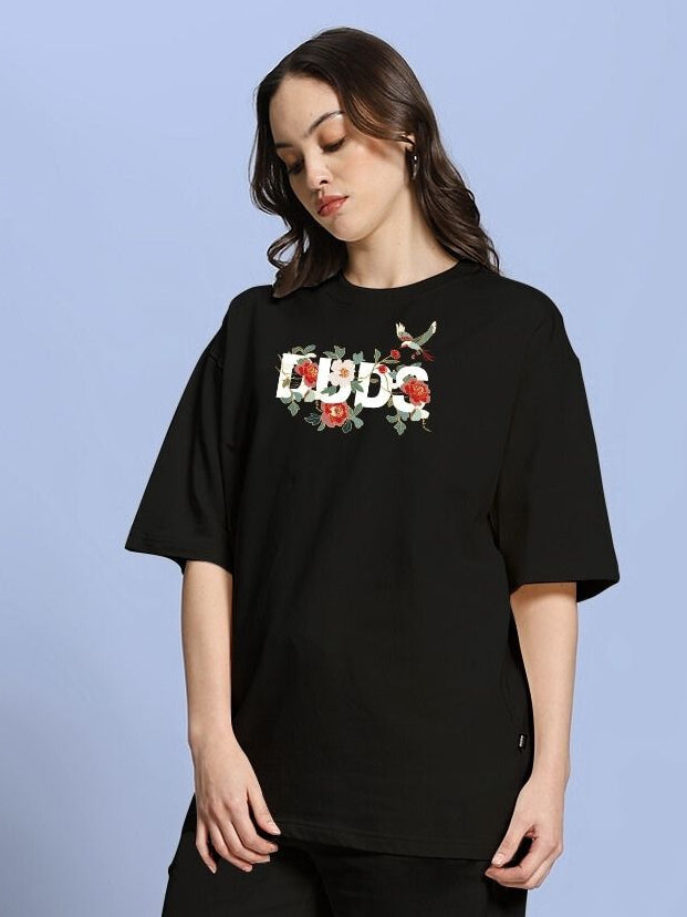 Woman wearing WOMEN'S ROSE CHARM OVER-SIZED T-SHIRT BLACK with floral design, hands to side, blue background