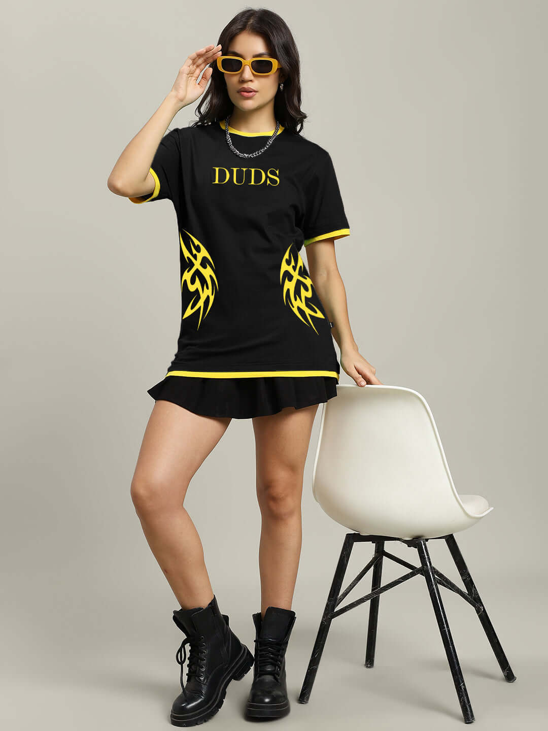 Woman wearing a black NIMO regular fit t-shirt with yellow flame design, paired with black boots and yellow sunglasses, standing by a white chair.