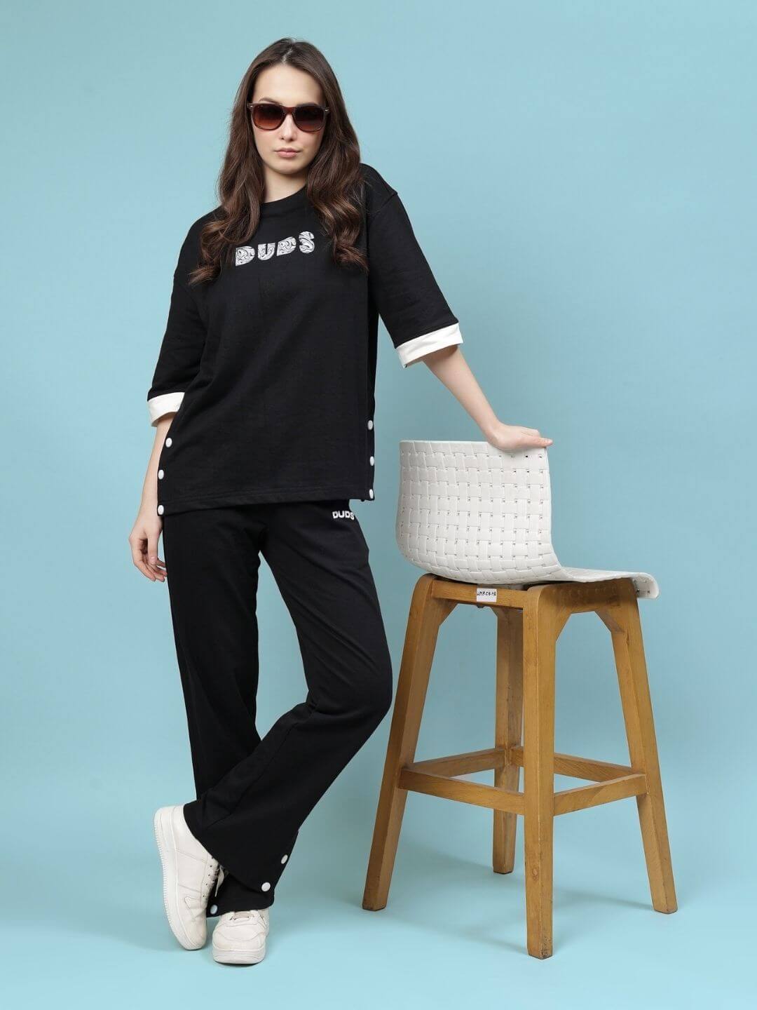 Woman wearing WOMEN'S WESENT RELAXED-FIT CO-ORD SET BLACK, styled with casual sneakers, posing by a wooden chair in a studio setting.