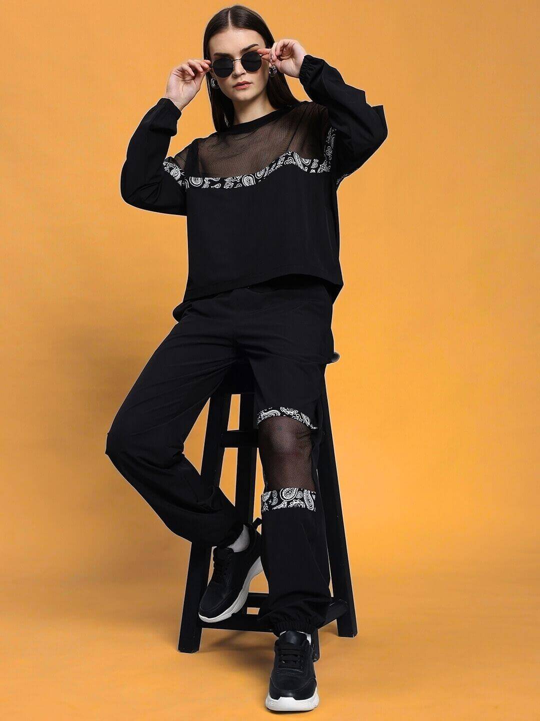 Woman wearing a black relaxed fit co-ord set with sheer details, sitting on a stool against an orange background.