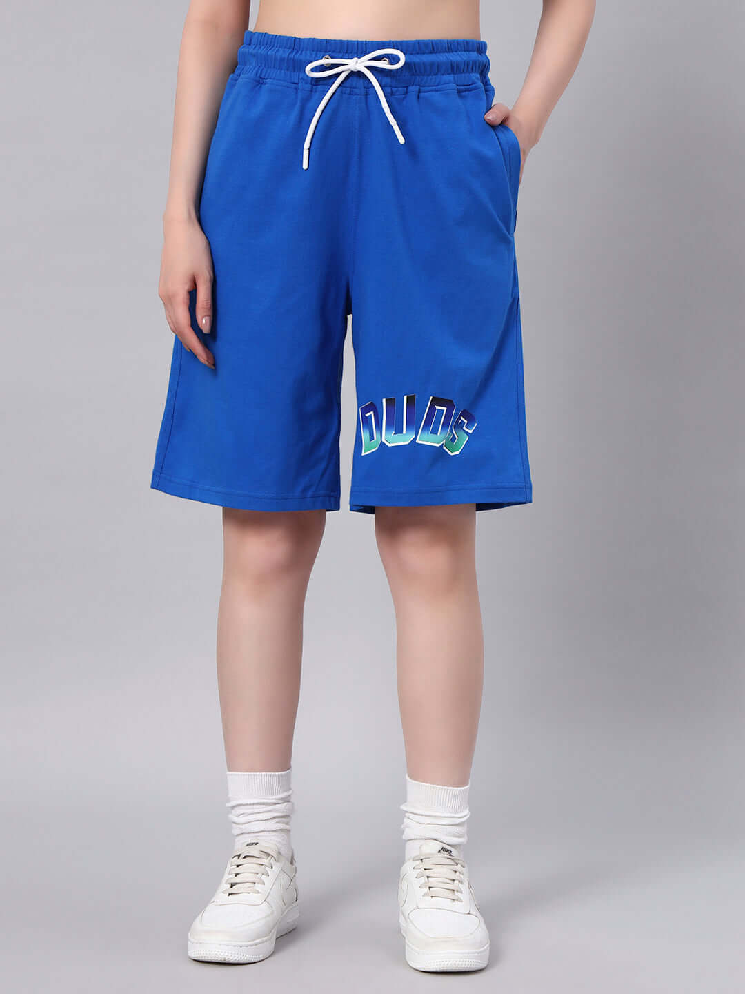 Model wearing blue shorts with 'DUDS' lettering, drawstring waist, front pockets, and white sneakers, showcasing stylish casual wear.