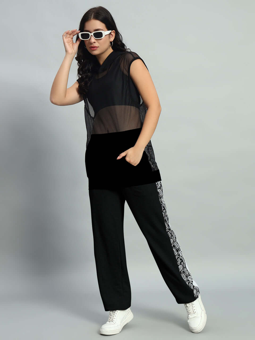 Woman in black pants with side patterns and white sneakers, wearing a sheer top and sunglasses, in a confident pose on a gray background.