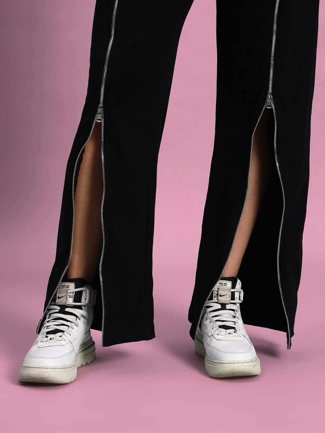 Black women's joggers with front zipper detail paired with white sneakers on a pink background.