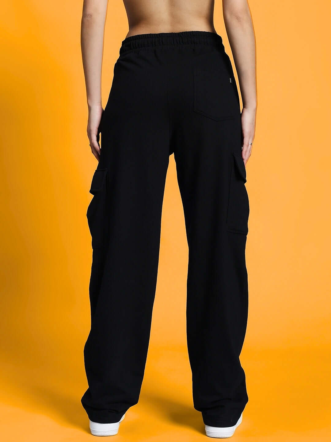 Back view of model wearing WOMEN'S BRUNO 5 POCKET CARGO PANT BLACK, showcasing utility style and feminine tailoring on orange background.