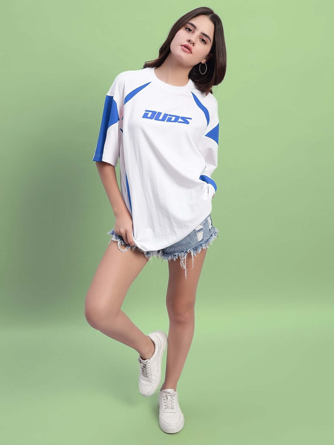 Model wearing Women's Candor Over-Sized T-Shirt in white and blue, crafted from premium cotton, styled with denim shorts, posing on green backdrop.
