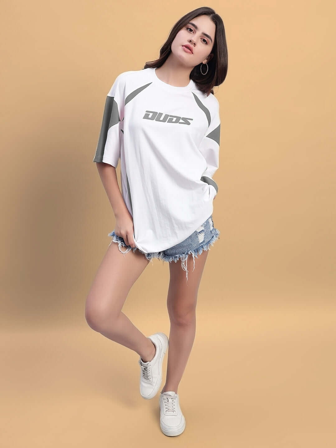 Model wearing women's oversized white grey t-shirt with bold design, paired with denim shorts and sneakers, showcasing casual fashion.