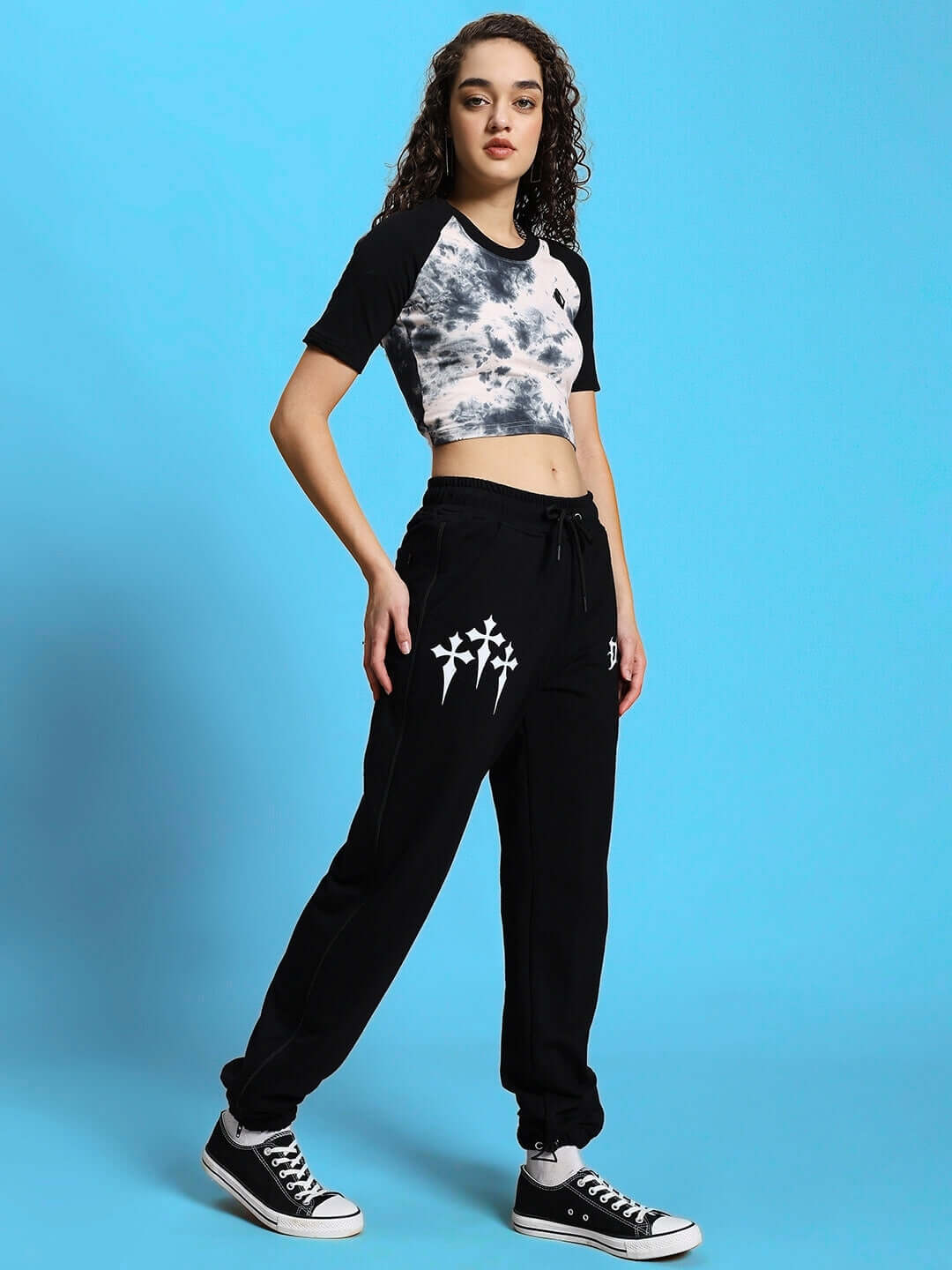 Model wearing trendy casual outfit with graphic tee and sneakers on blue background.