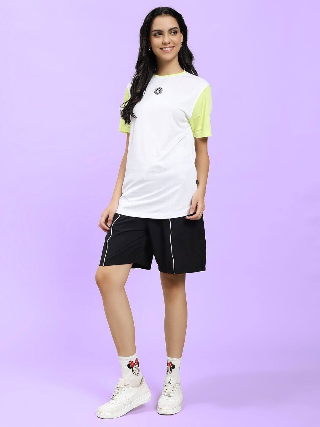 Woman wearing WOMEN CHAMPION ZONE REGULAR FIT CO-ORD WHITE NEON BLACK outfit with neon sleeve and black shorts on purple background