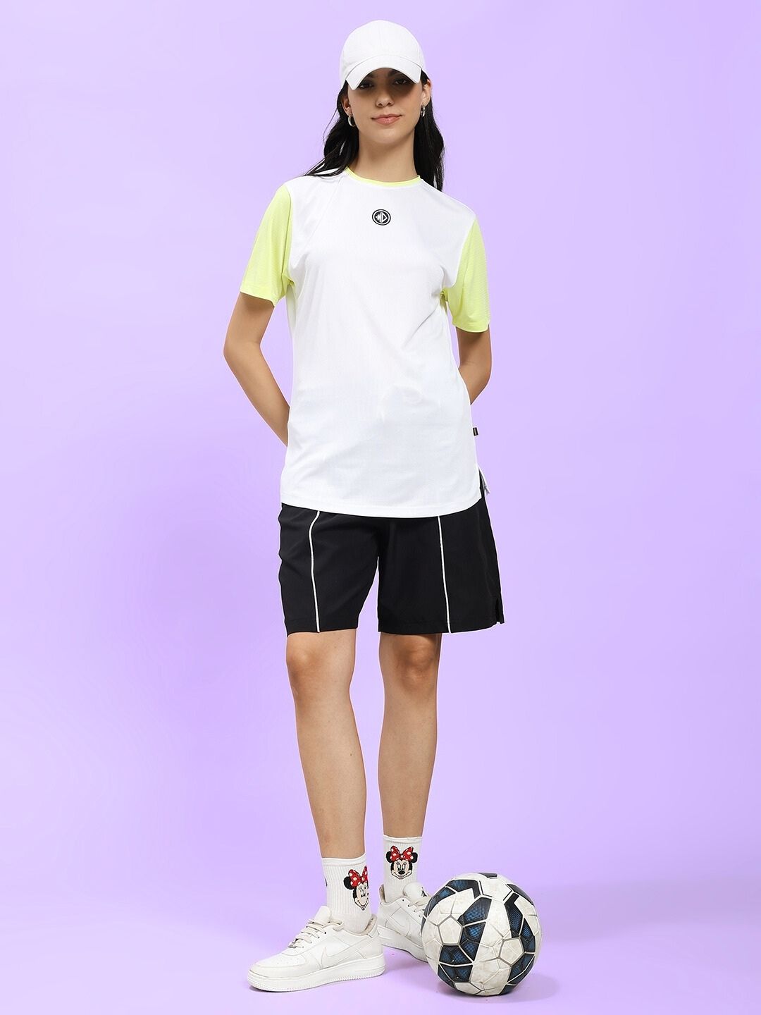 Woman wearing WOMEN CHAMPION ZONE REGULAR FIT T-SHIRT WHITE NEON-GREEN, paired with black shorts, standing next to a soccer ball