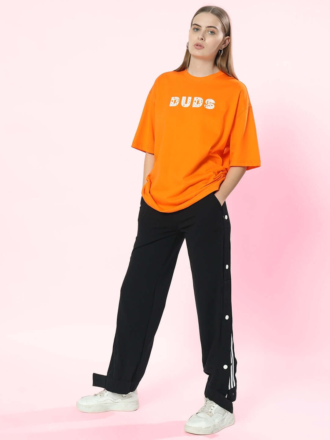 Model wearing black women's hip hop joggers by DUDS, paired with an orange DUDS t-shirt and white sneakers, standing against a pink background.