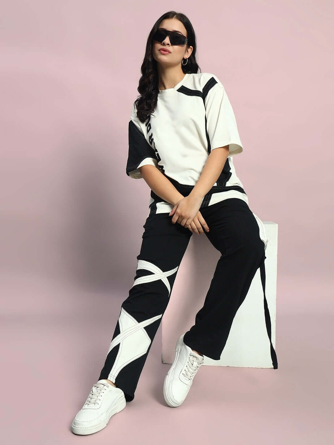 Woman wearing off white-black oversized co-ord set with relaxed fit joggers, styled with sunglasses, sitting against a pastel background.