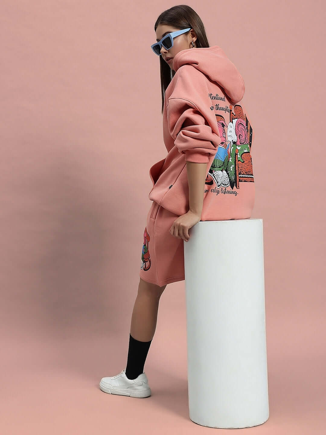 Woman wearing WOMEN DELIBERATE FLEECE SHORTS PEACH with matching hoodie and sunglasses, posing on a pink background for a stylish look.
