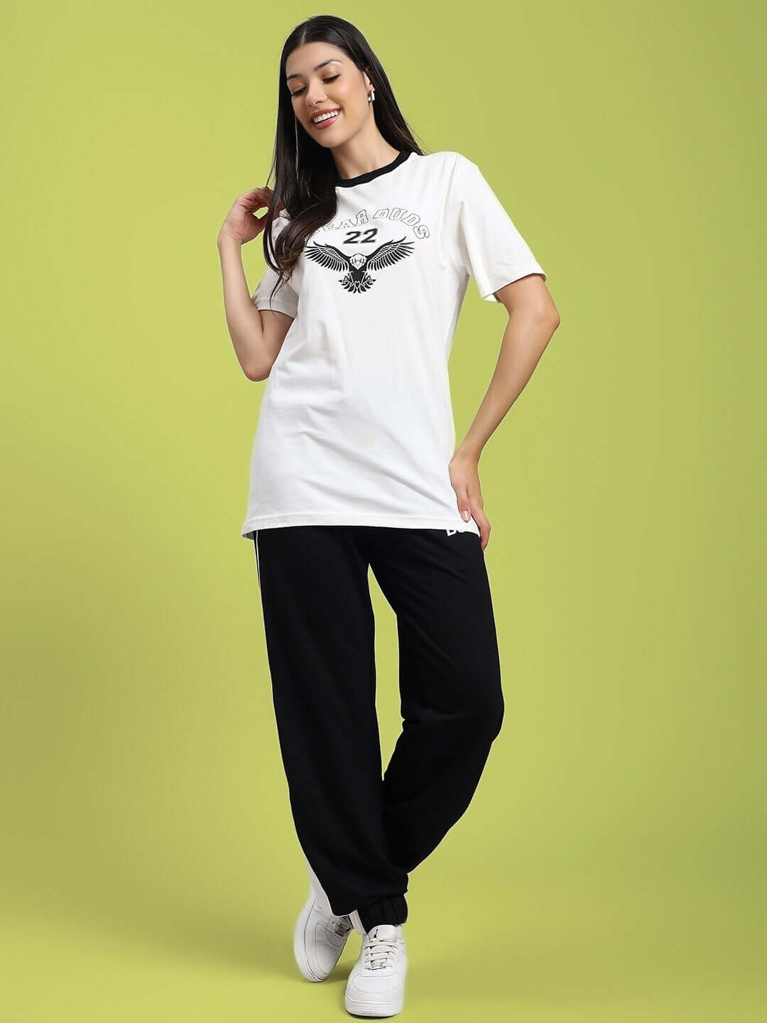 Woman wearing WOMENS DENVER RELAXED FIT JOGGERS BLACK, with a white graphic t-shirt and sneakers, standing against a green background.