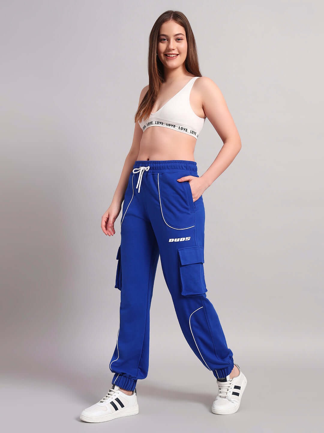 Model wearing Women's Devono Relaxed-Fit Joggers in blue with cargo pockets, paired with a white bralette and sneakers.