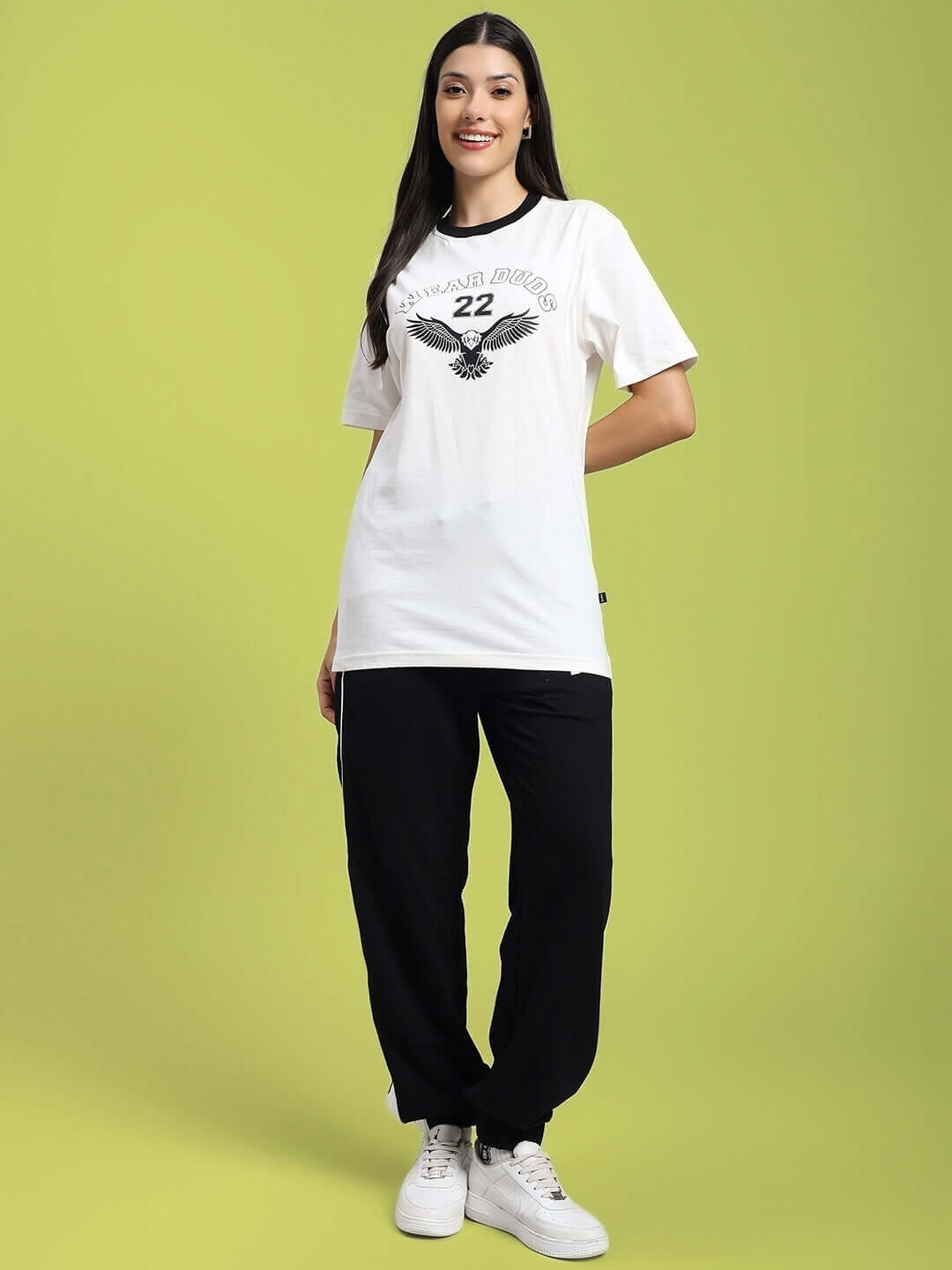Woman wearing White Eagle Wings T-Shirt, black pants, and white sneakers on green background.
