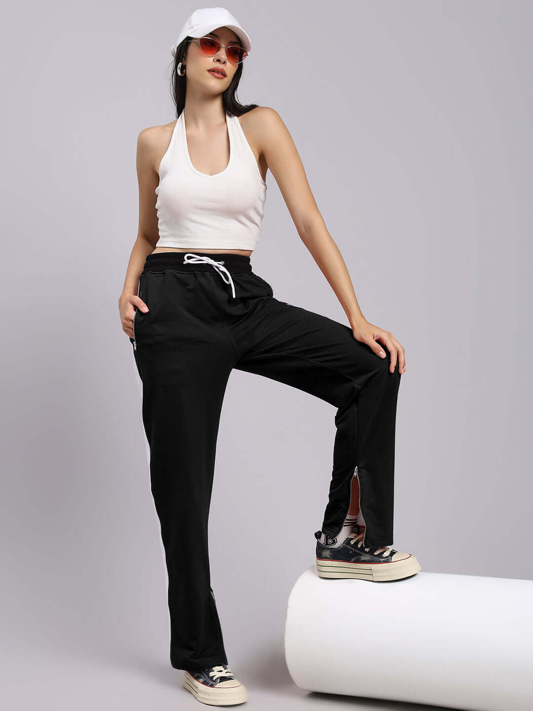 Woman wearing black relaxed-fit joggers and a white tank top, paired with casual sneakers and a cap, stands confidently against a light backdrop.