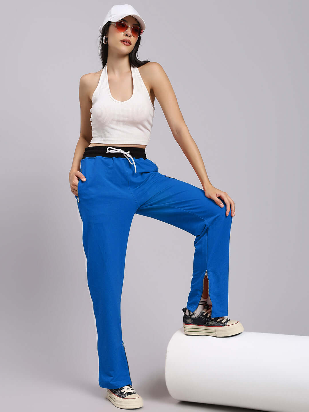 Model wearing women's elegant relaxed-fit joggers in blue, paired with a white tank top and cap, showcasing casual sporty style.