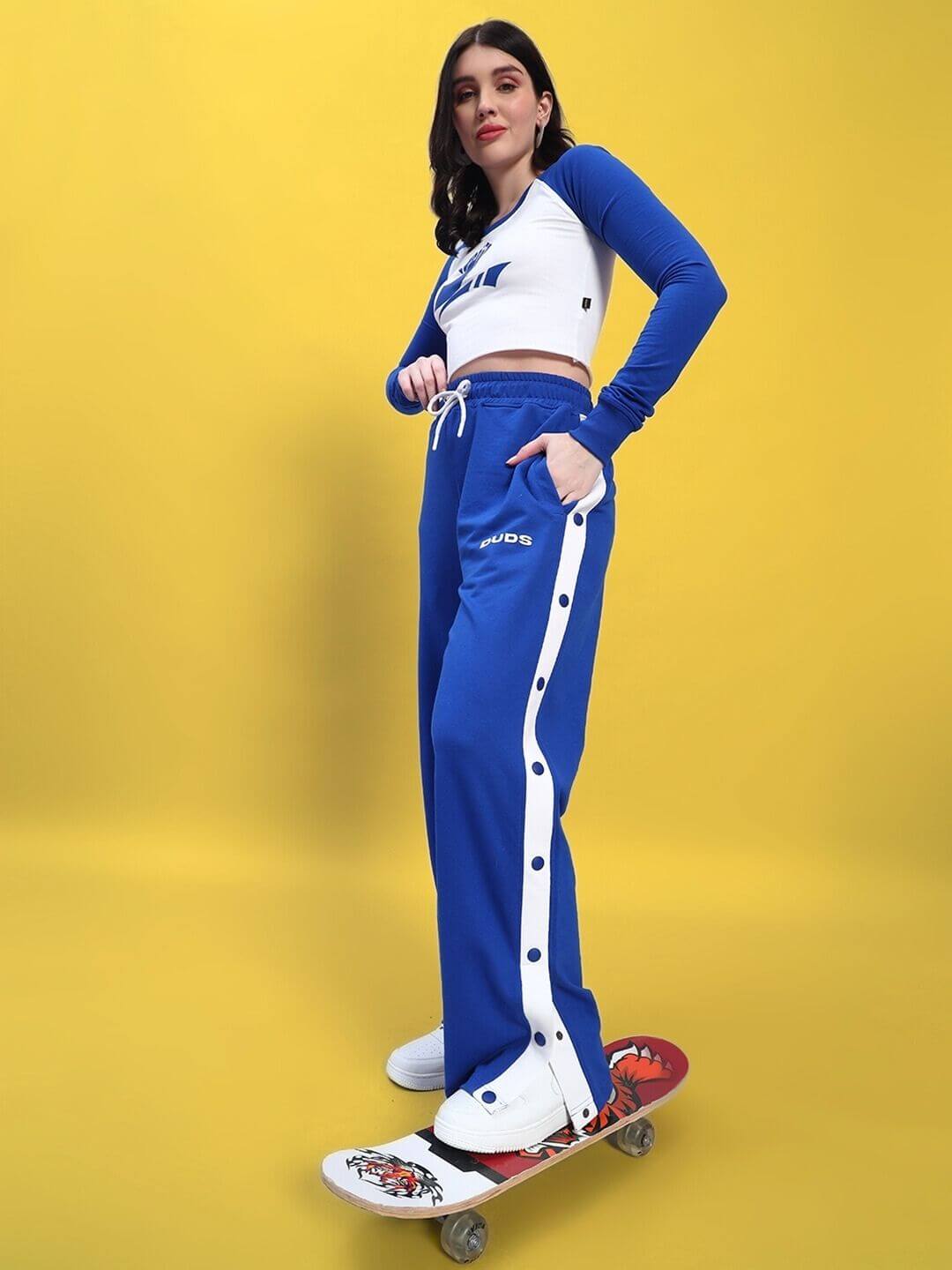 Model wearing WOMEN'S EZEL CO-ORD SET ROYAL BLUE-WHITE, standing on skateboard against yellow background, showcasing stylish athletic look.