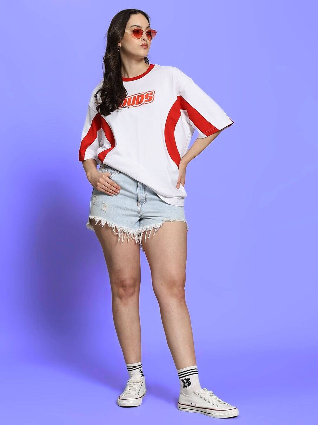Woman wearing oversized white-red Women's Ezel T-shirt and denim shorts, standing confidently against a purple background.