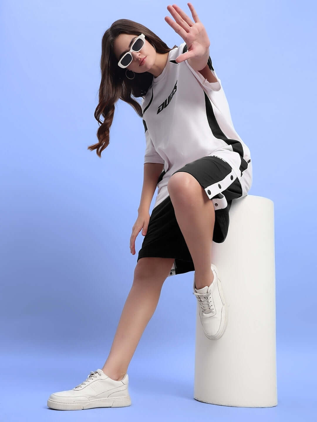 Woman in black shorts and white top posing on a pedestal against a blue background, wearing sunglasses and sneakers.