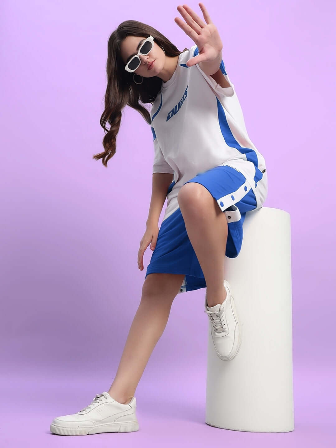 Model wearing blue and white casual attire, sitting on a white stool with a pink background, showcasing trendy women's fashion.