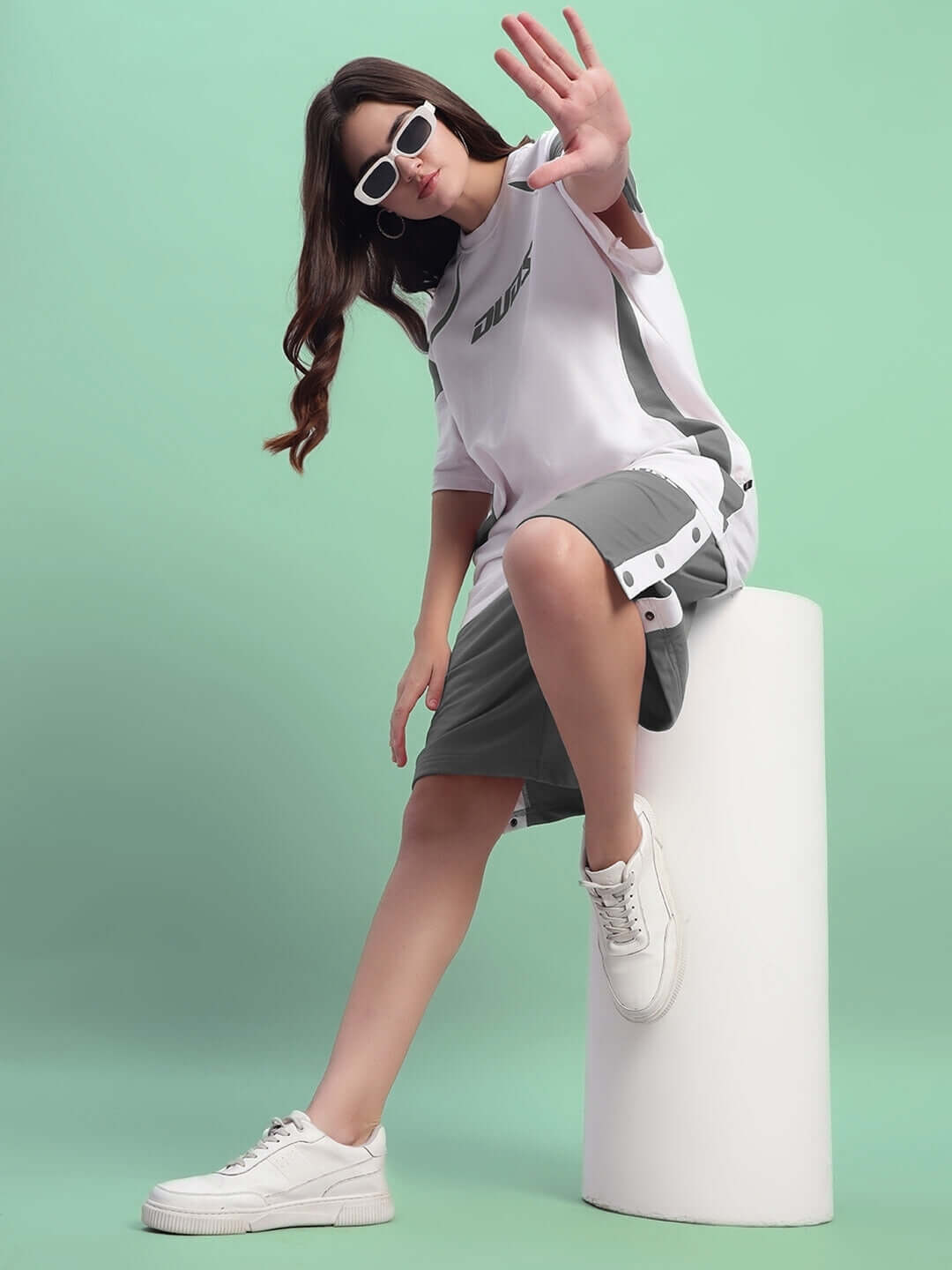 Woman wearing DUDS WOMEN'S EZEL REGULAR FIT SHORTS in grey, sitting on white stool against mint green backdrop, stylish casual look.