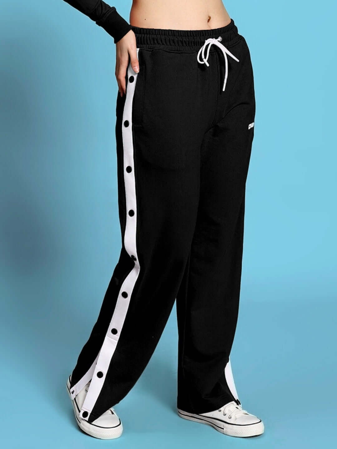 Woman wearing black relaxed fit joggers with side button detail and white sneakers on a blue background.