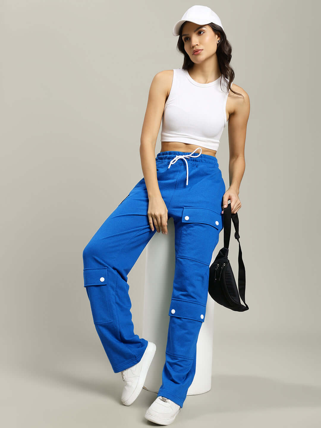 Woman wearing blue flap pocket baggy joggers paired with a white crop top and cap, holding a black bag.