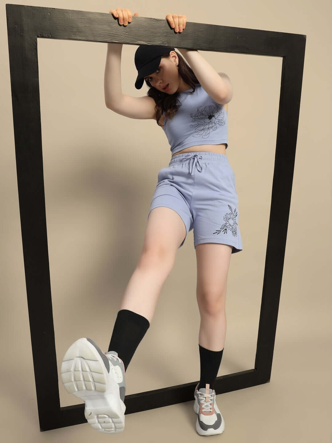 Woman in steel blue co-ord set with floral print, wearing sneakers and a cap, posing with a large wooden frame.