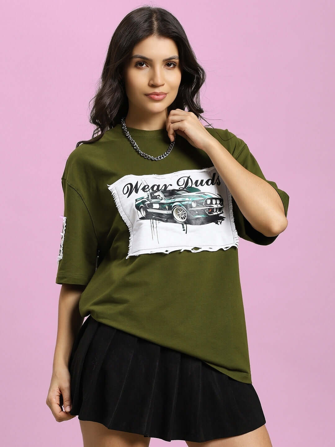 Woman wearing olive green oversized Ford Mustang t-shirt with black skirt, standing in front of pink background.