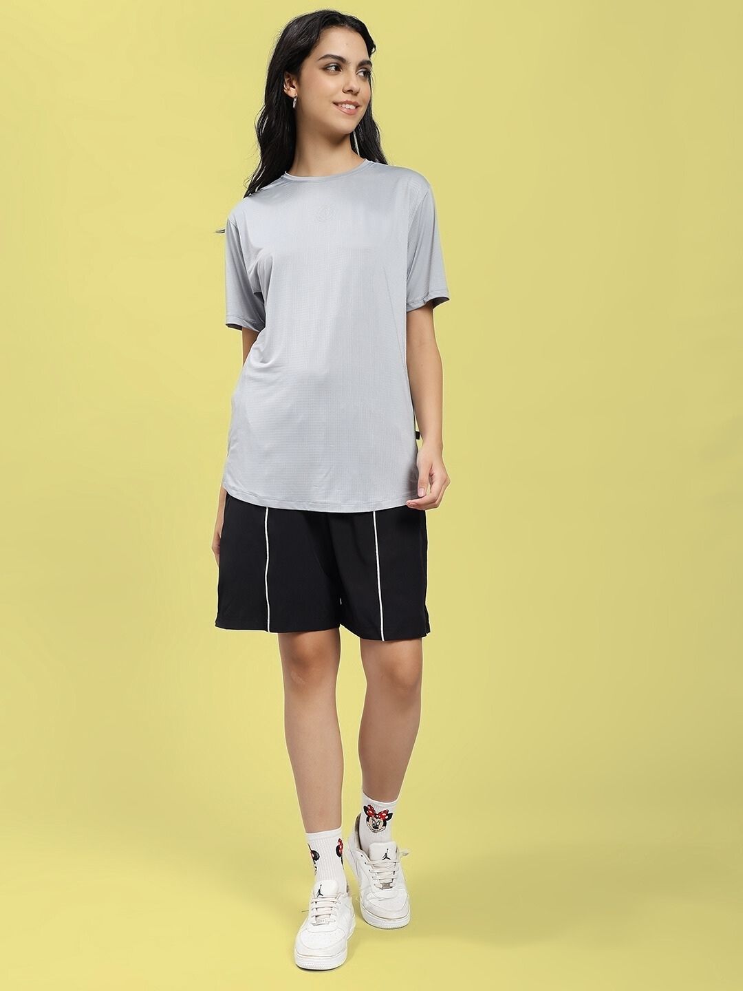 Model wearing WOMEN JET RUN REGULAR FIT T-SHIRT GREY paired with black shorts against a yellow background.