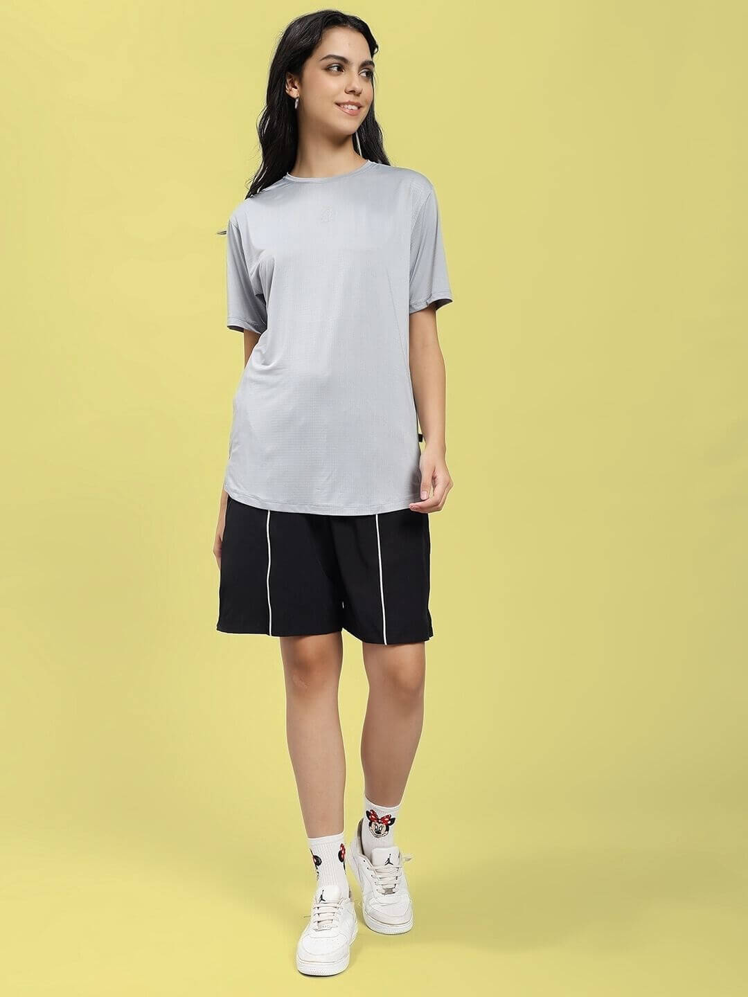 Woman wearing WOMEN JET RUN REGULAR FIT T-SHIRT GREY paired with black shorts and white sneakers on a yellow background.