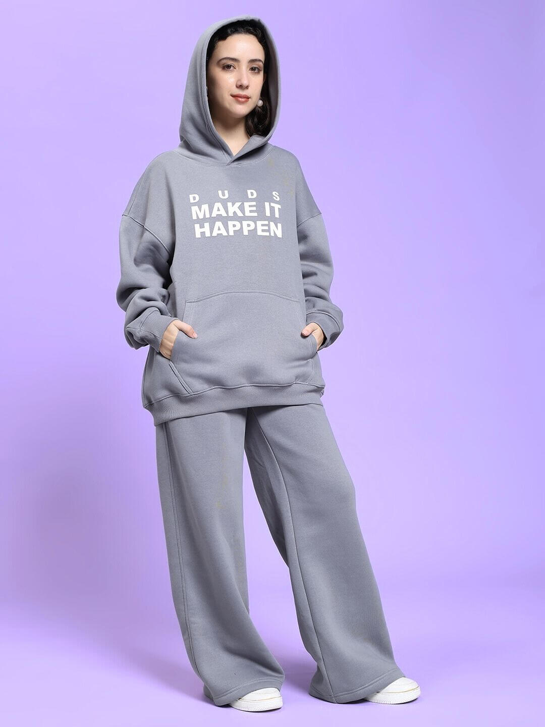 Woman wearing the WOMEN MAKE IT HAPPEN FLEECE CO-ORD SET GREY, featuring a cozy hoodie and wide-leg pants, on a purple background.