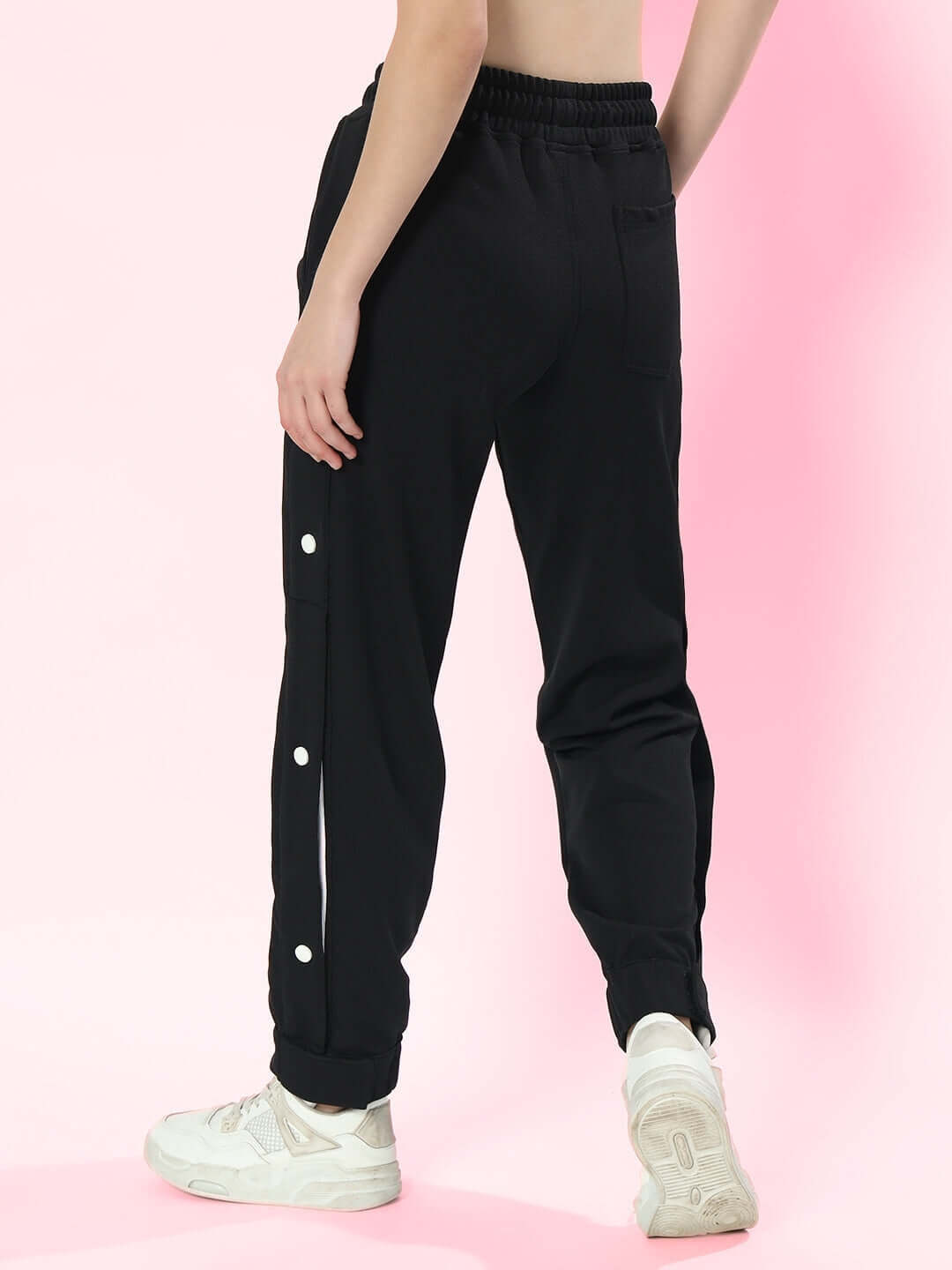 Woman wearing black hip hop joggers with side buttons, styled with white sneakers, against a pink background.