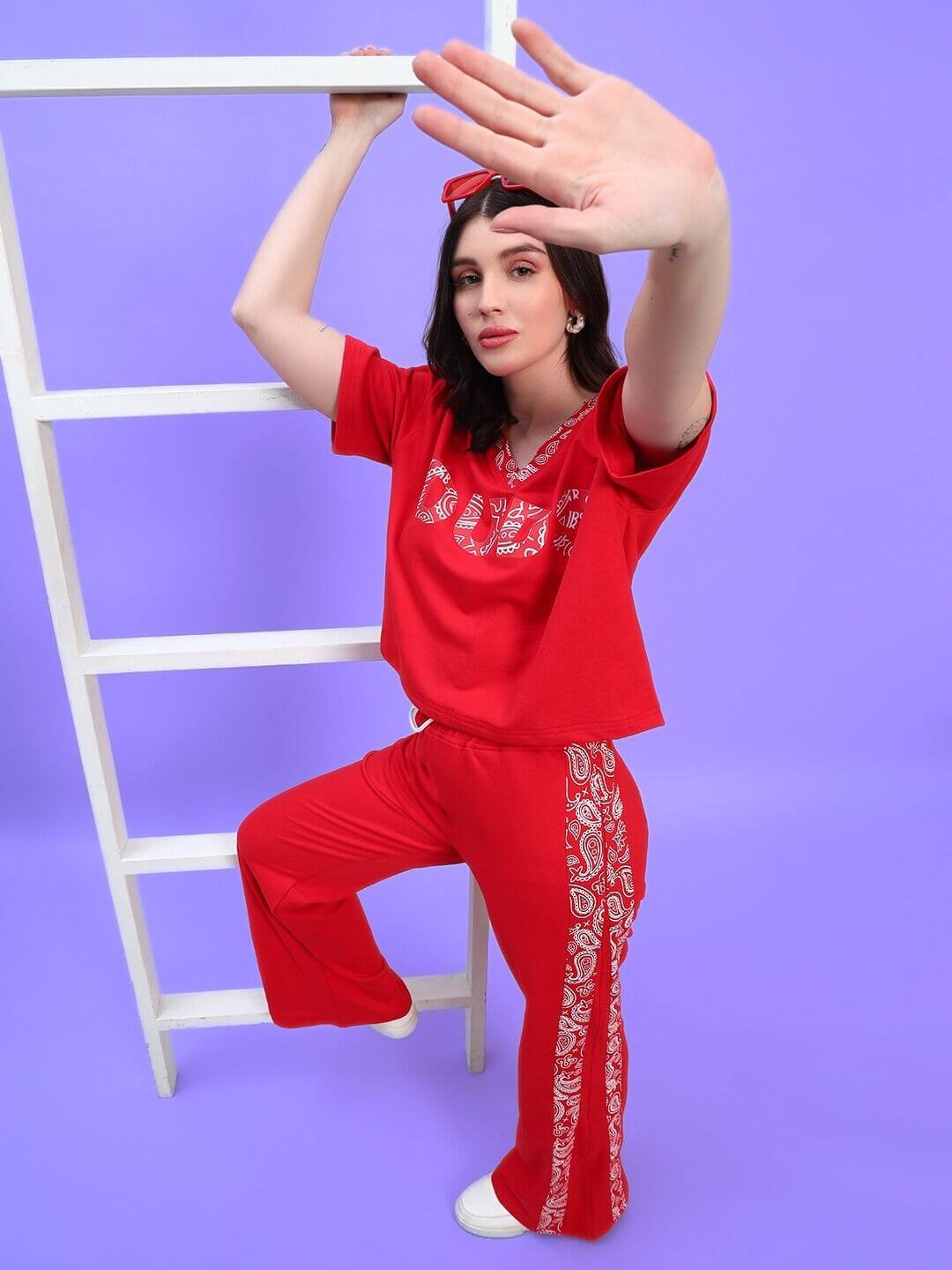 Woman posing in red streetwear outfit with graphic design, leaning on a white ladder, against a purple backdrop.