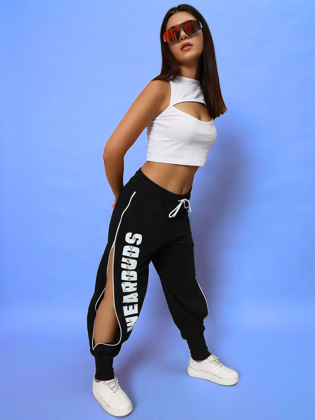 Model wearing black women's joggers with side slits, white sneakers, white crop top, and sunglasses against a blue background.