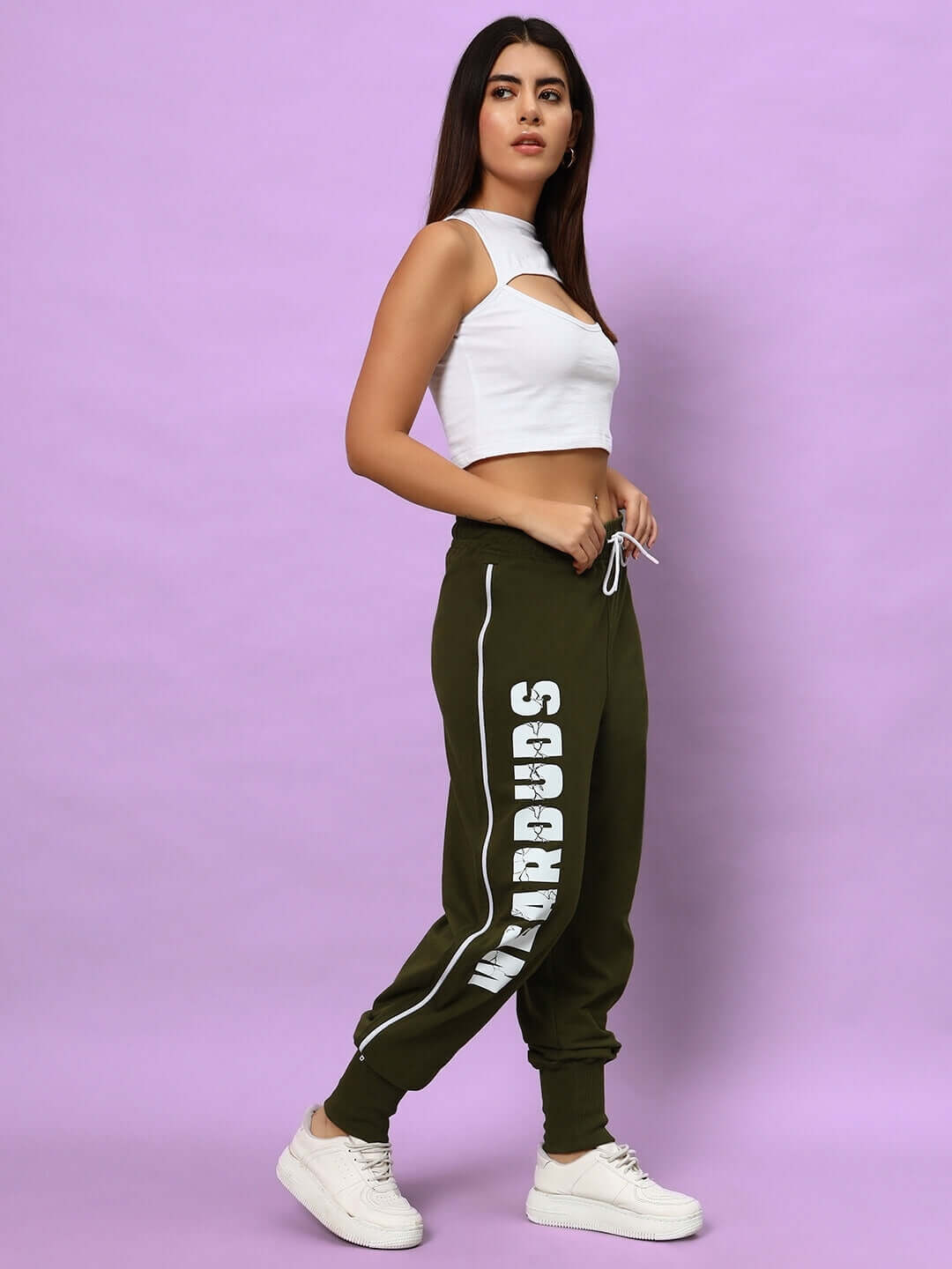 Woman wearing olive green Hunter joggers by DUDS, featuring white side lettering, paired with a white crop top and sneakers on a purple background.