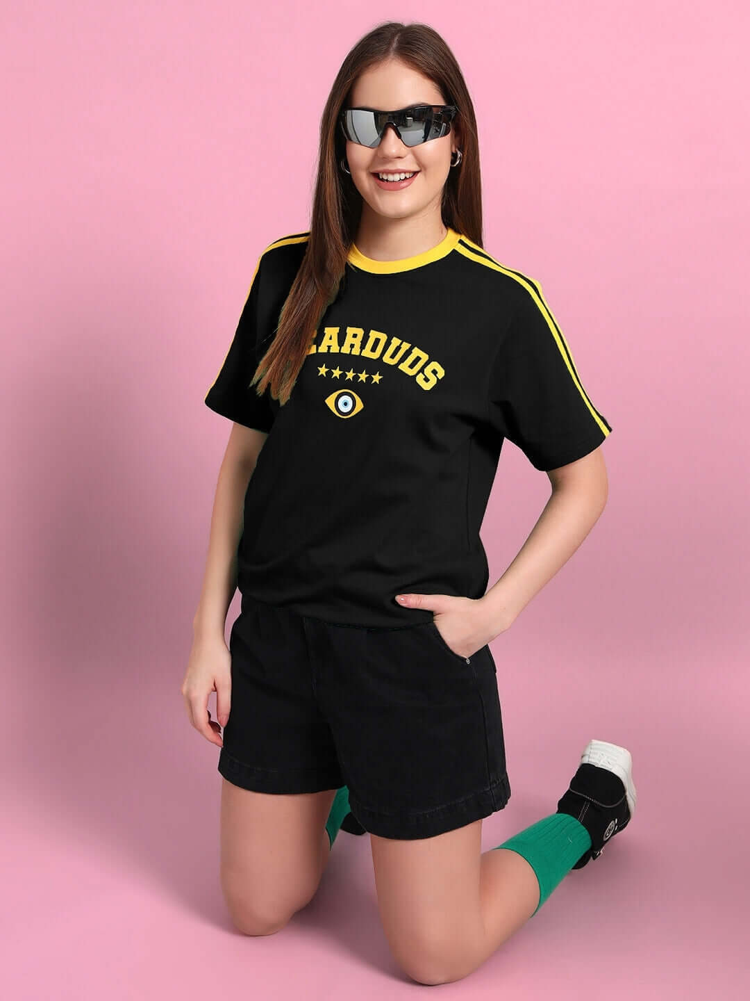 Woman wearing black t-shirt with yellow accents, black shorts, and sunglasses against a pink background.