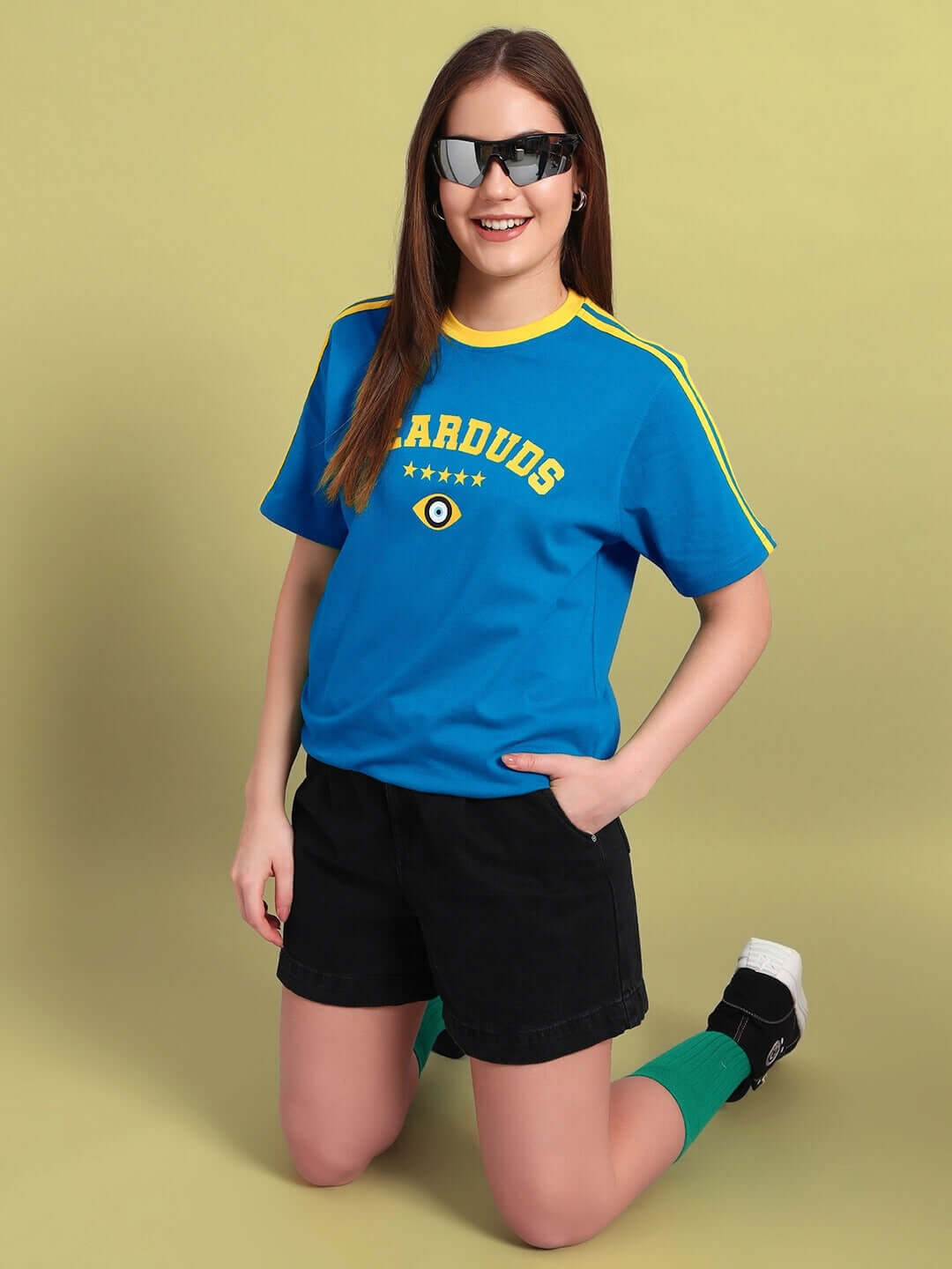 Woman wearing a blue Irish regular fit t-shirt with 'Stardust' text, styled with black shorts and sunglasses, kneeling on a light background.