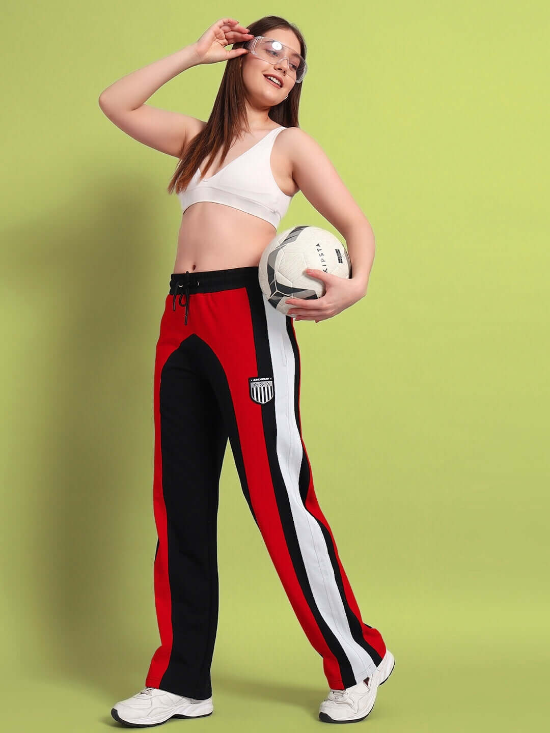 Woman wearing Jamison color block relaxed-fit joggers in black and red, holding a soccer ball, smiling against a lime green background.