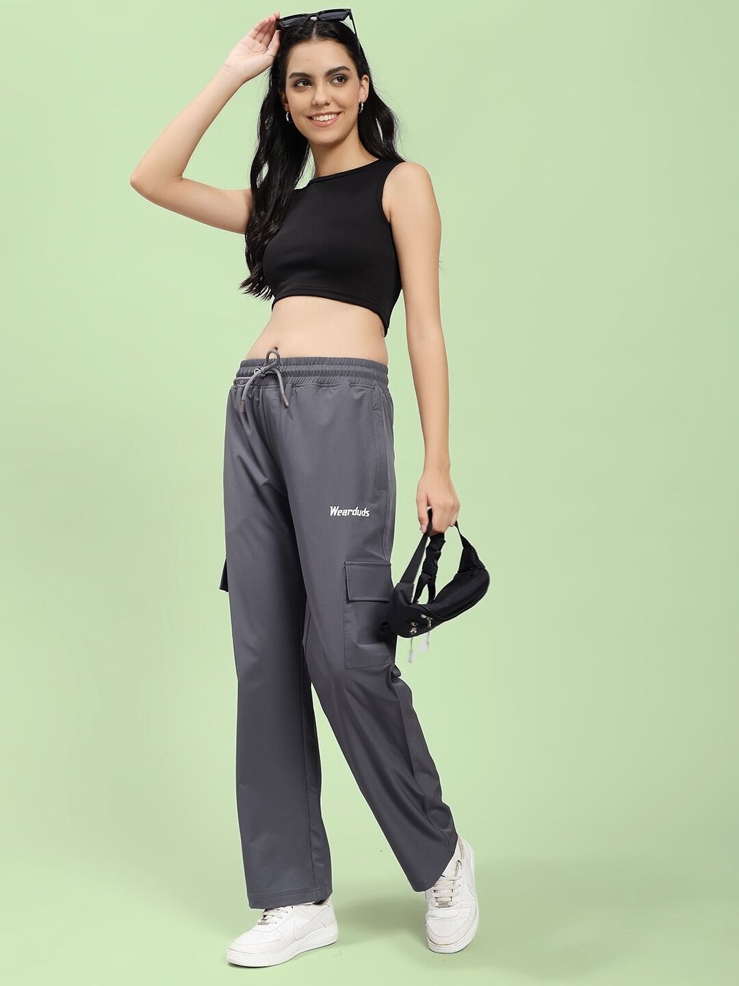 Woman wearing WOMEN'S JIMMY RELAXED FIT MULTI POCKET JOGGER DARK GREY with black top and white sneakers, holding sunglasses.