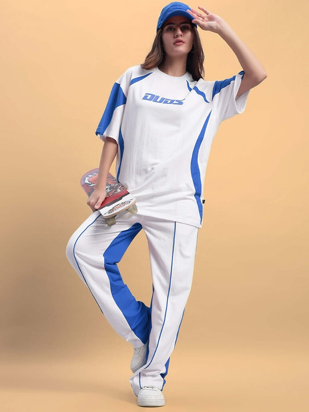Woman wearing white and blue relaxed fit joggers and top, holding skateboard. Casual fashion by DUDS. Order now for custom fit options.