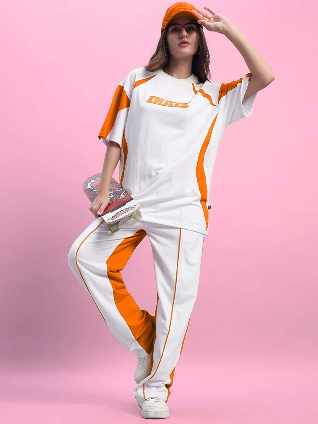 Model wearing white orange DUDS joggers with relaxed fit, holding a skateboard, against pink background.