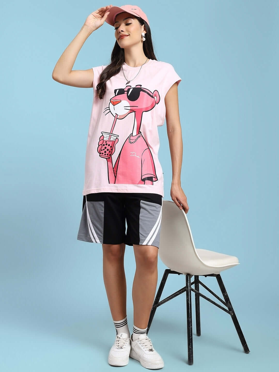 Woman in a pink cartoon t-shirt with black shorts, standing next to a white chair against a blue background.