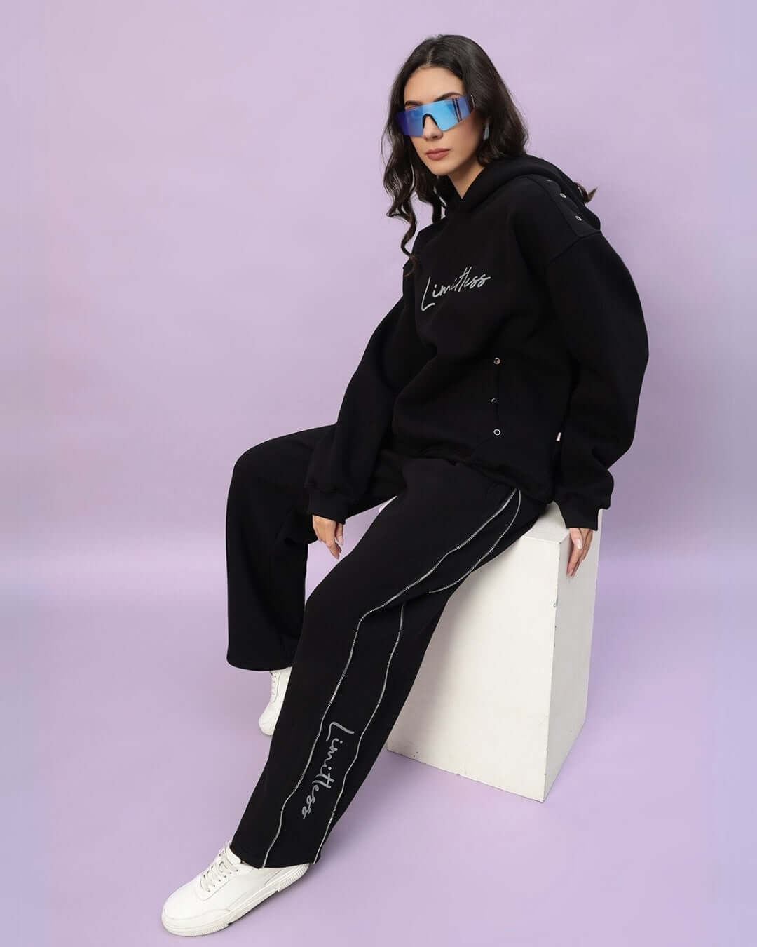 Woman wearing a black Limitless Fleece Reflector Hoodie and pants with sunglasses, posing against a purple background.